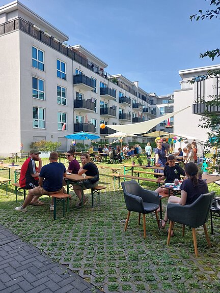 Menschen sitzen bei sonnigem Wetter auf Bänken und Stühlen im Innenhof eines modernen Wohnkomplexes. Über ihnen spenden Sonnensegel Schatten. Es herrscht eine lebhafte, entspannte Stimmung.
