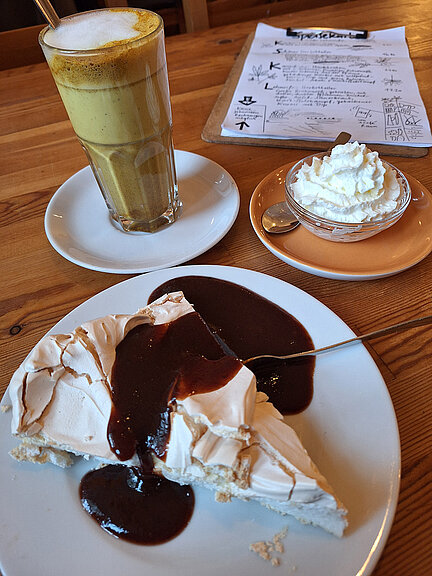 A warm drink, a piece of cake and a small bowl of whipped cream can be seen on a café table. 
