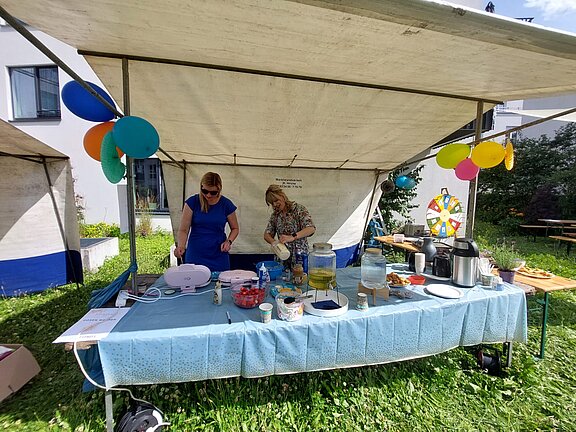 Zwei Frauen bereiten unter einem festlich dekorierten Pavillon Essen und Getränke vor. Auf dem Tisch stehen verschiedene Speisen, Getränke und Küchenutensilien. Im Hintergrund ist ein Glücksrad zu sehen.
