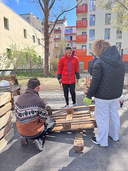 Drei Personen arbeiten im Freien gemeinsam an Holzpaletten. Eine Person kniet, zwei steht und halten Werkzeug. Im Hintergrund sind Wohnhäuser, Bäume und blauer Himmel zu sehen. Es wirkt wie ein Gemeinschaftsprojekt im Stadtviertel.