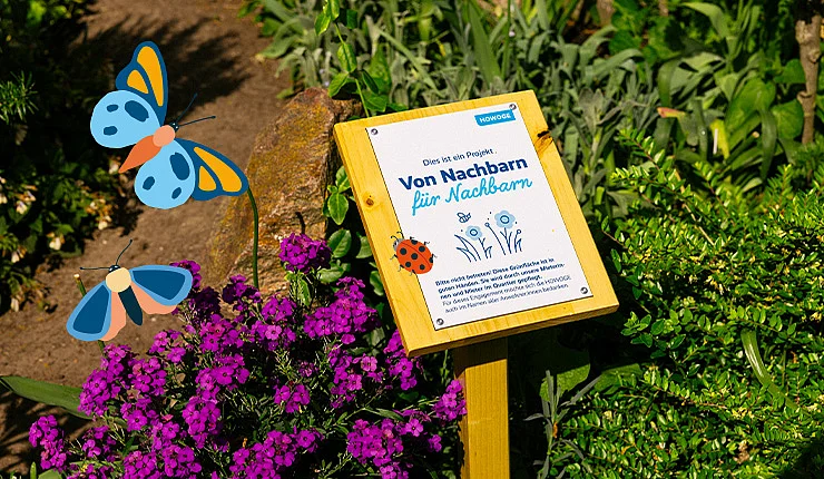Eine Infotafel vor einem Blumenbeet. Auf der Tafel steht "Von Nachbarn für Nachbarn".