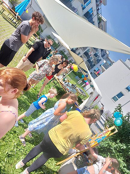Menschen feiern bei sonnigem Wetter ein Fest im Freien. Unter Sonnensegeln stehen Kinder und Erwachsene zusammen, es gibt bunte Luftballons und Spiele, vermutlich in einer Wohnanlage oder Nachbarschaft.