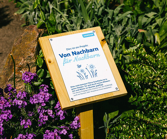 Eine Infotafel vor einem Blumenbeet. Auf der Tafel steht "Von Nachbarn für Nachbarn".