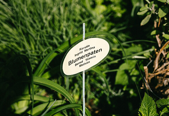 Ein Schild in einem Blumenbeet. Auf dem Schild steht "Blumenpaten".