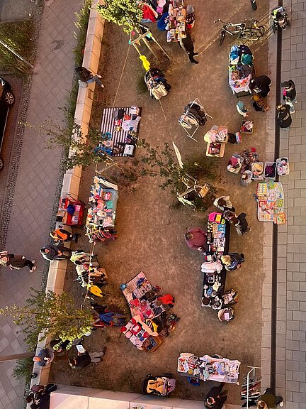 Blick von oben auf einen belebten Flohmarkt im Freien: Mehrere Stände mit bunten Decken und Waren, Menschen schlendern und stöbern bei sonnigem Wetter auf einem schmalen Grünstreifen zwischen Gehwegen.