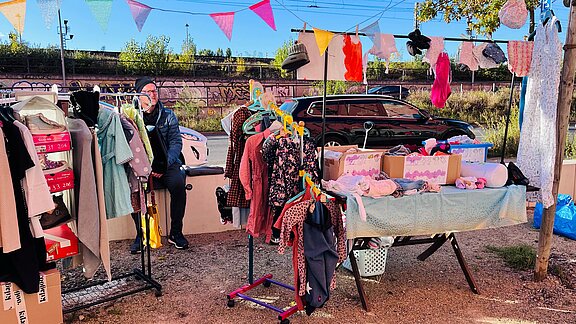 Bunter Flohmarktstand im Freien mit Kleidung auf Kleiderständern, Accessoires und Kartons auf einem Tisch. Eine Person sitzt im Hintergrund, bunte Wimpel hängen über dem Stand, im Hintergrund parkt ein Auto.