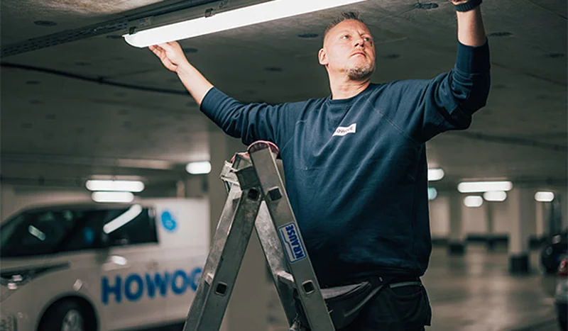 Ein Mann steht in einem Parkhaus auf einer Leiter und bring eine neue Leuchtstoffröhre an der Decke an. Im Hintergrund steht ein Auto, auf welchem Werbung der Howoge zu sehen ist.