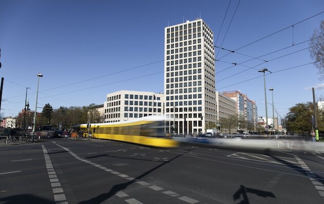 Bild – Blick auf die Kreuzung am Stefan Heym Platz. Eine Straßenbahn und Autos fahren vor dem neugebauten Turm vorbei.