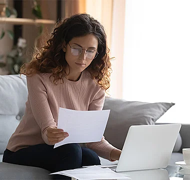 Frau mit lockigen Haaren und Brille sitzt auf einem Sofa, betrachtet konzentriert ein Dokument und arbeitet an einem Laptop. Neben ihr steht eine Tasse auf dem Tisch. Die Szene wirkt ruhig und fokussiert im Homeoffice.
