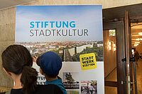 Eine Frau steht mit ihrem Kind vor einer Wandtafel über die Stiftung Stadtkultur