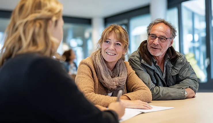 Ein Mann und eine Frau sitzen mit einer Frau an einem Tisch in einem öffentlichen Gebäude und lassen sich beraten. Sie lächeln, es herrscht eine freundliche Atmosphäre. 