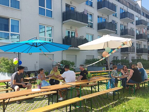 Menschen sitzen an Biertischen im Innenhof eines modernen Wohnhauses. Bunte Sonnenschirme und Girlanden sorgen für eine gemütliche Atmosphäre bei Sonnenschein.