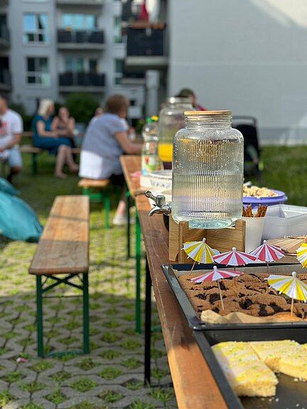 Ein bunt gedeckter Tisch mit Getränken und Kuchen im Freien, im Vordergrund Gläser und Gebäck mit kleinen Sonnenschirmchen. Im Hintergrund sitzen Menschen auf Bänken bei einem gemütlichen Treffen.