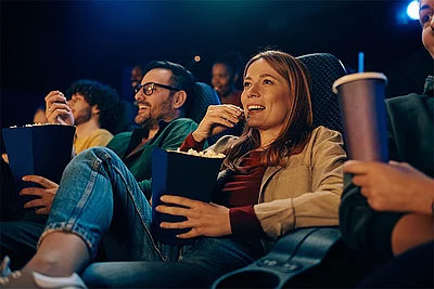 Mehrere Menschen sitzen in einem Kinosaal und schauen nach vorn. Im Vordergrund sitzt eine Frau, die einen Popcorneimer in der Hand hält und Popcorn isst.