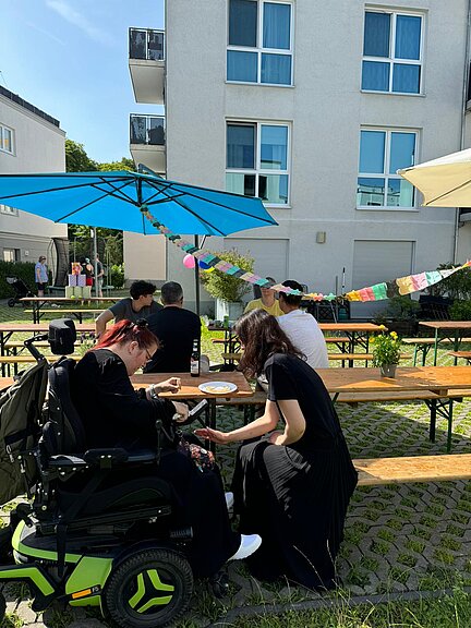 Menschen sitzen an Biertischen im Innenhof eines Wohnhauses. Es ist sonnig, bunte Girlanden und Sonnenschirme schmücken den Platz. Eine Frau im Rollstuhl und eine andere Person unterhalten sich im Vordergrund. Sommerliche, entspannte Stimmung.