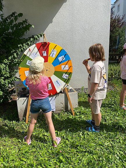 Zwei Kinder stehen bei sonnigem Wetter im Gras vor einem bunten Glücksrad, das mit Bildern und Farben gestaltet ist. Eines der Kinder dreht gerade am Rad, das andere schaut gespannt zu und isst ein Eis.