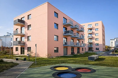Rosa farbige Fassade von dem Neubau, davor Grünwiese mit Spielplatz