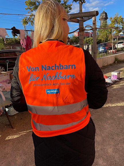 Eine Person trägt eine orange Warnweste mit der Aufschrift „Von Nachbarn für Nachbarn – ehrenamtlich aktiv bei der HOWOGE“. Sie steht im Freien bei sonnigem Wetter, im Hintergrund sind Verkaufsstände zu sehen.