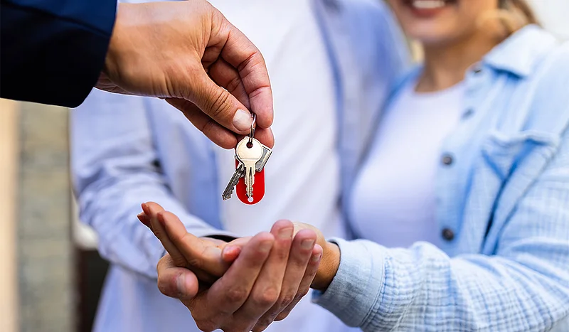 Ein Paar erhält symbolisch den Schlüssel zu seinem neuen Zuhause – eine Hand übergibt ihnen ein Schlüsselbund, während sie strahlend die Hände entgegenstrecken. Ein Moment voller Vorfreude und Neuanfang.