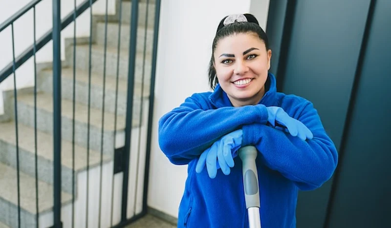 Eine dunkelhaarige Frau mit Handschuhen und einem blauen Pullover lehnt an einem Besen