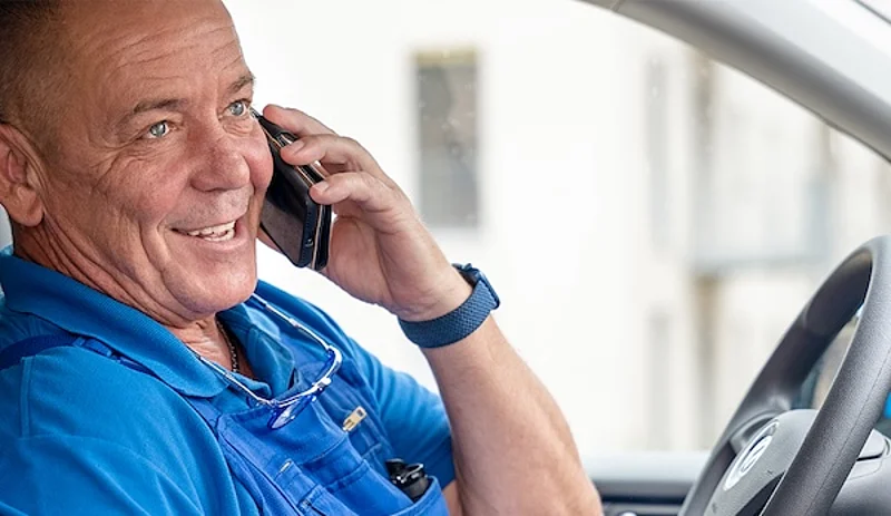 Ein Mann in einem blauen T-Shirt und blauer Latzhose mit HOWOGE Logo sitzt telefonierend im Auto und lacht