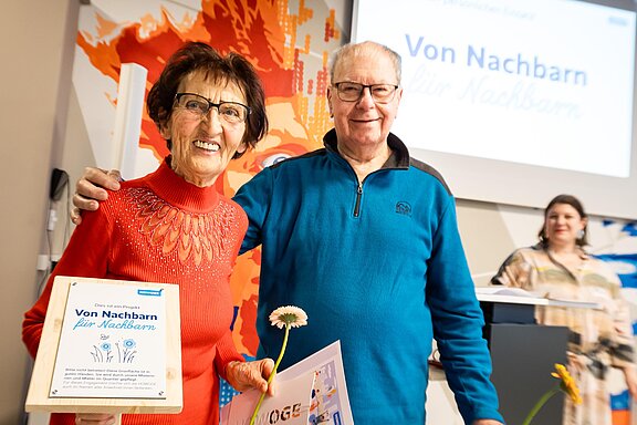 Zwei ältere Menschen lächeln in die Kamera und halten eine Urkunde und Blume in der Hand – sie wurden bei einer Veranstaltung mit dem Titel „Von Nachbarn für Nachbarn“ für ihr Engagement ausgezeichnet.