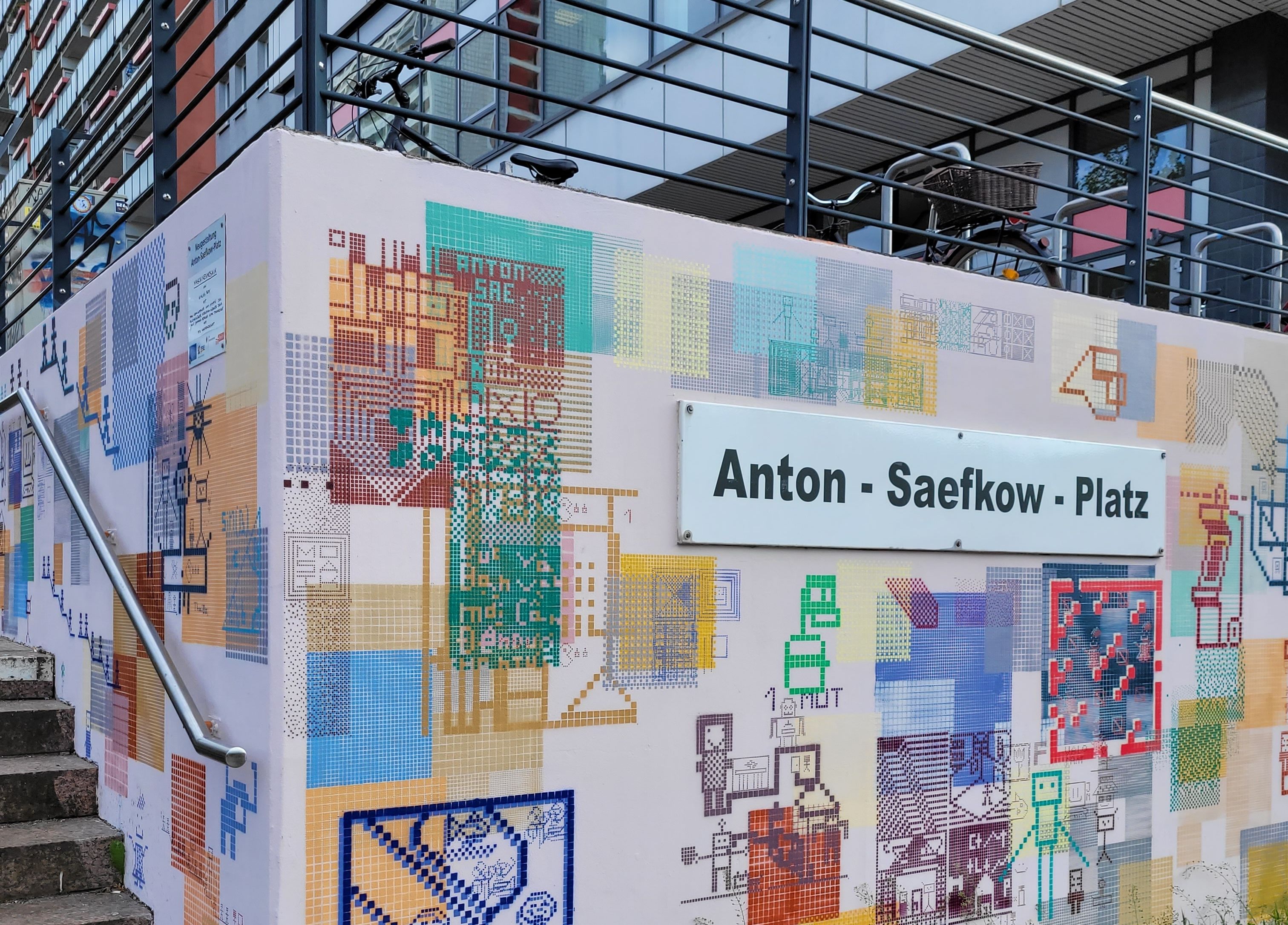 Bunte Wand mit Pixelkunst-Grafiken und einem Straßenschild „Anton-Saefkow-Platz“ an einem modernen Gebäude mit Treppe und Geländer im Vordergrund.