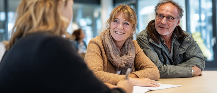 Ein Mann und eine Frau sitzen mit einer Frau an einem Tisch in einem öffentlichen Gebäude und lassen sich beraten. Sie lächeln, es herrscht eine freundliche Atmosphäre. 
